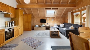 A wooden interior with a kitchen, dining area, and living room, featuring wooden beams, a flat-screen TV, and a sofa.