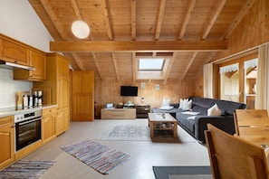 A wooden interior with a kitchen, dining area, and living room, featuring wooden beams, a flat-screen TV, and a sofa.