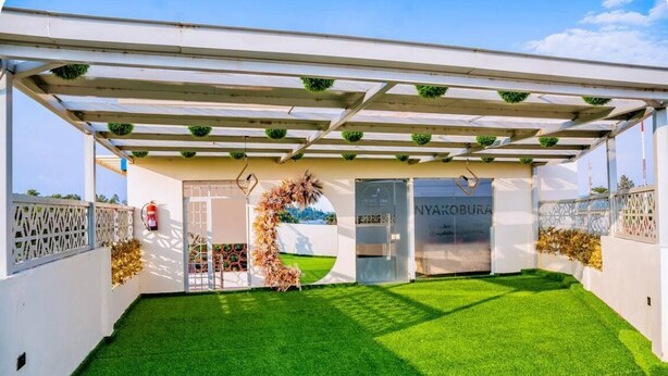 Un espace extérieur abrité avec une pelouse artificielle, un bâtiment blanc doté d’une porte en verre et une enseigne sur laquelle on peut lire « NYAKOBURA ».