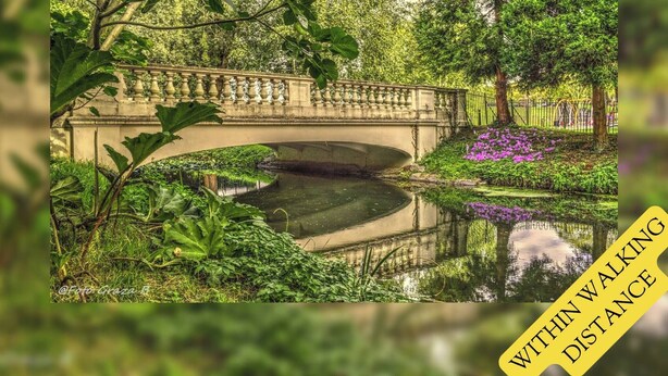 Een stenen brug over stil water, omringd door weelderige begroeiing en bloeiende planten.