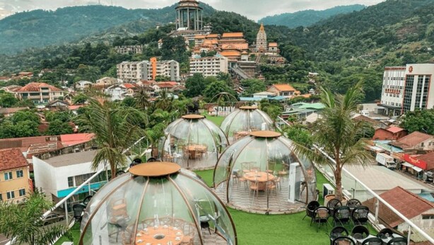 Una terraza en la azotea con cúpulas transparentes, mesas y sillas, con vistas a un paisaje urbano que incluye edificios y un templo al fondo.