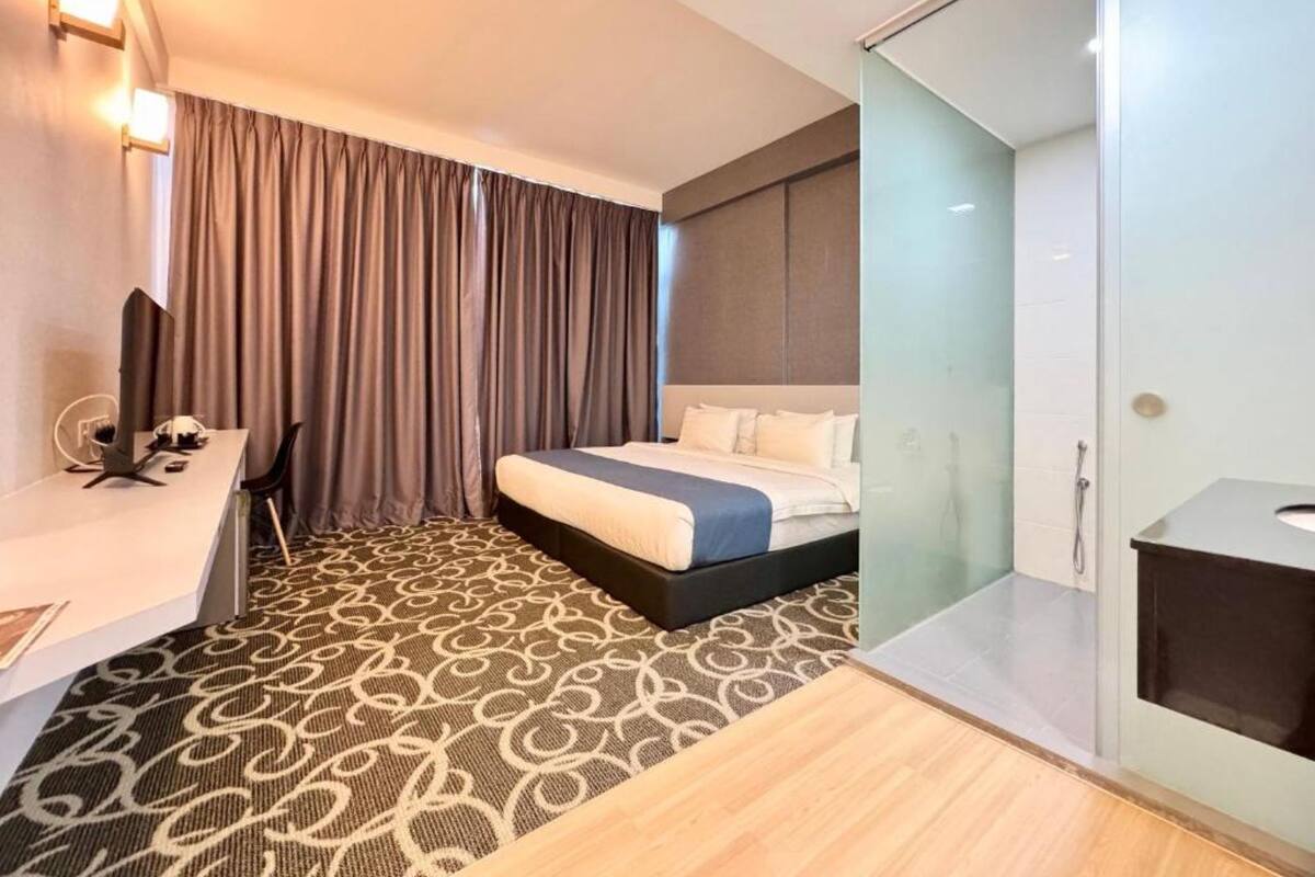 A hotel room with a bed, a desk, a chair, a glass partition, and a shower area.