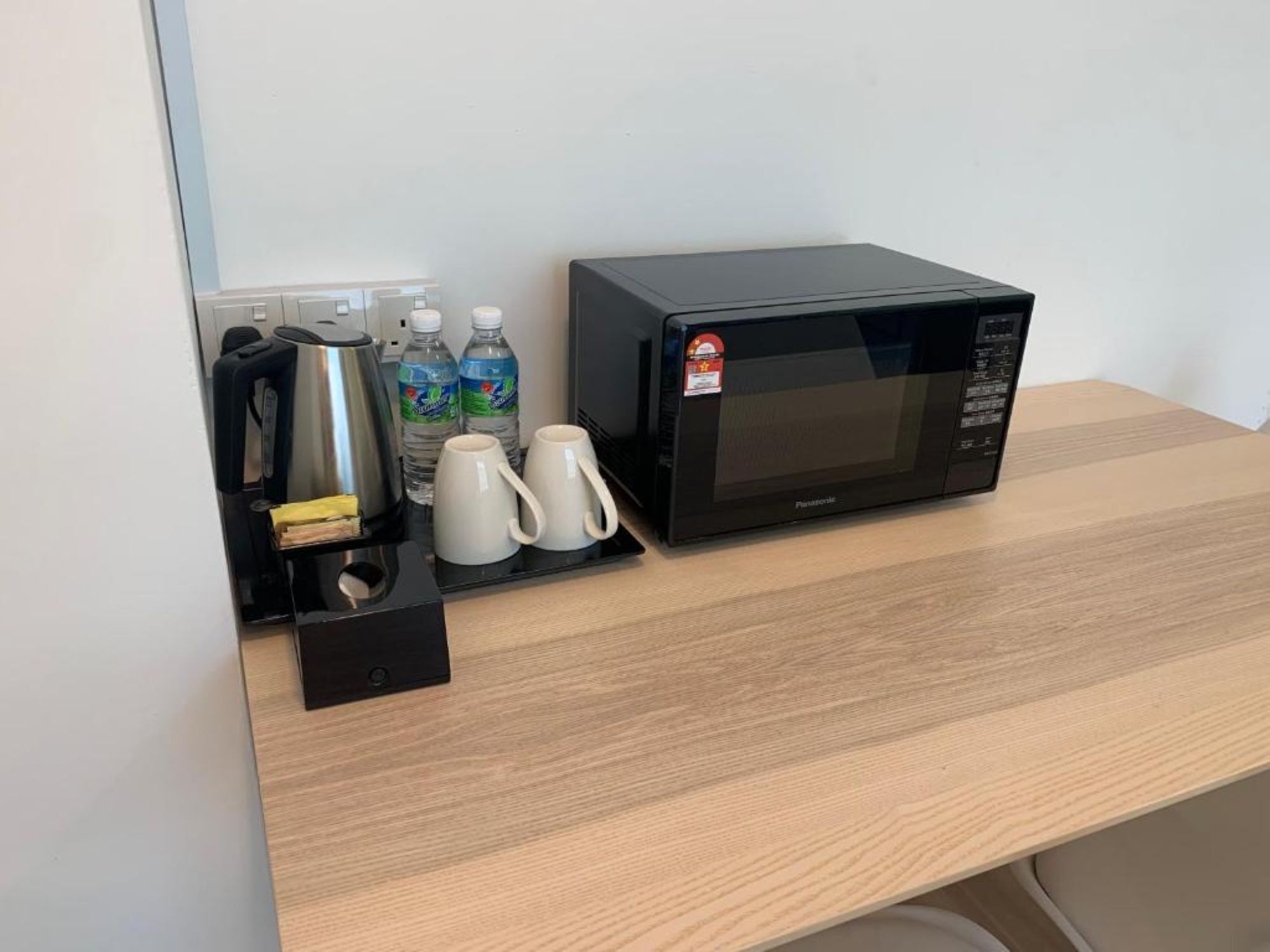 A microwave on a wooden countertop with a coffee maker, two water bottles, and two white mugs.