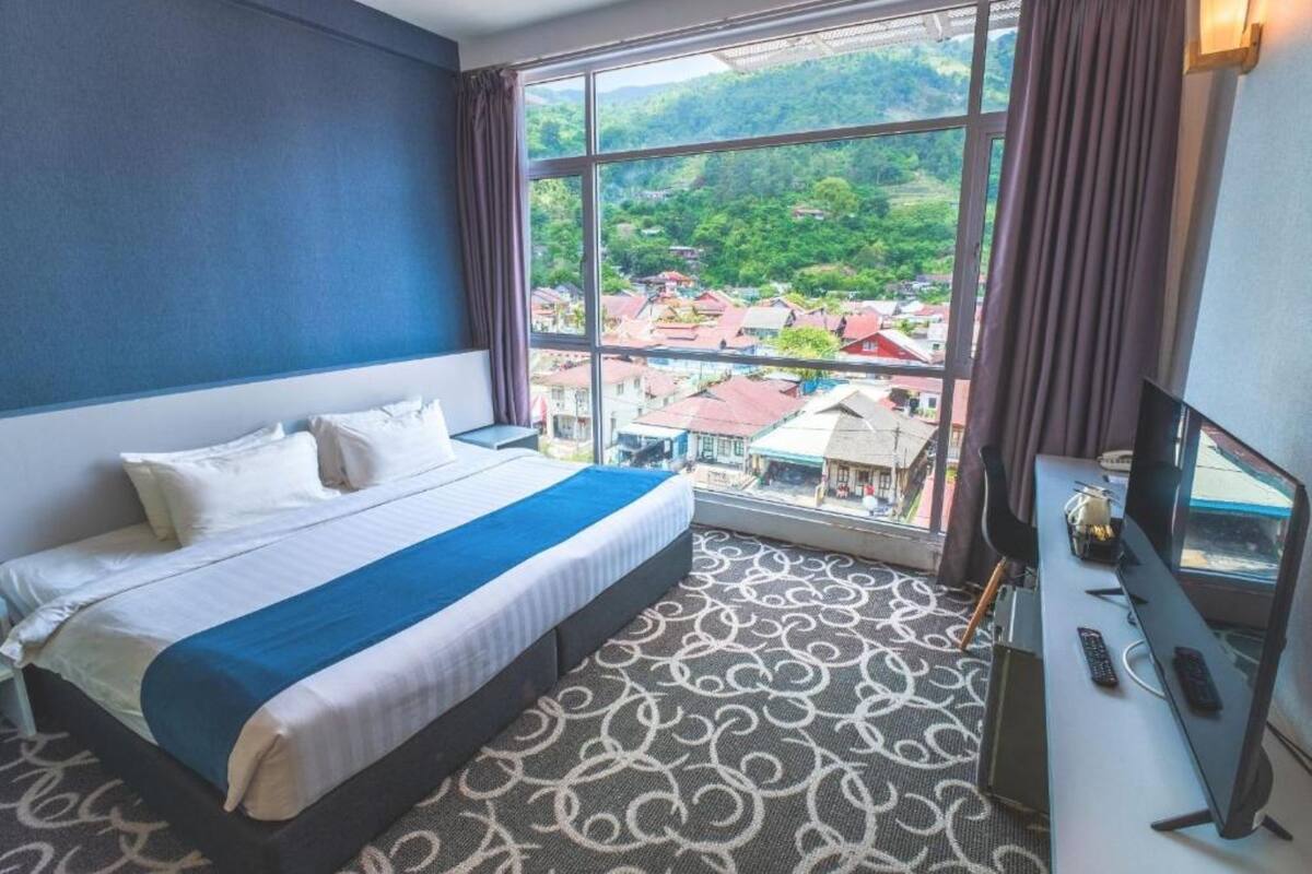 A hotel room with a large bed, a desk with a computer, and a view of a town through large windows.