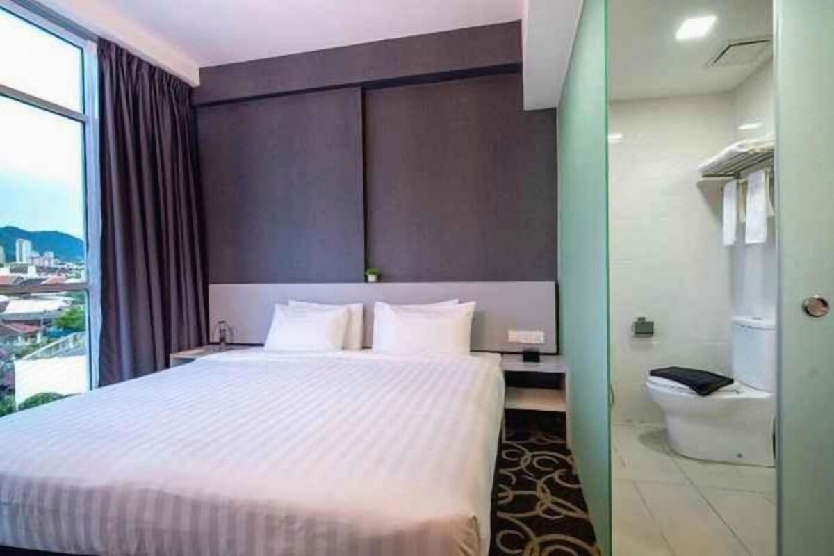 Une chambre d’hôtel avec un grand lit, une table de chevet, une commode, une fenêtre avec des rideaux et une salle de bain équipée de toilettes et d’une douche.