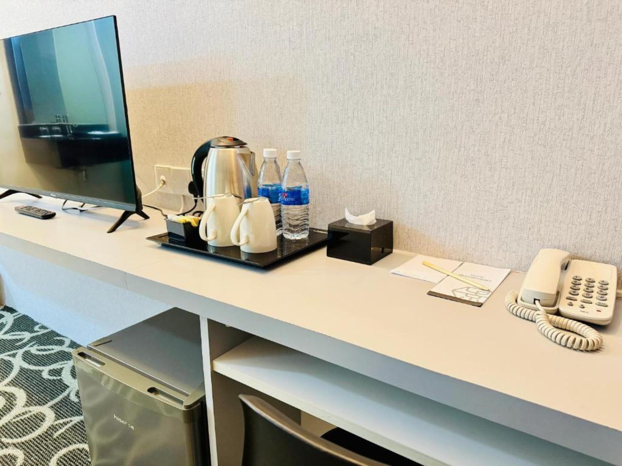 Un mostrador de recepción de hotel con una televisión de pantalla plana, una cafetera, botellas de agua, una caja de pañuelos, un teléfono y un cubo de basura.