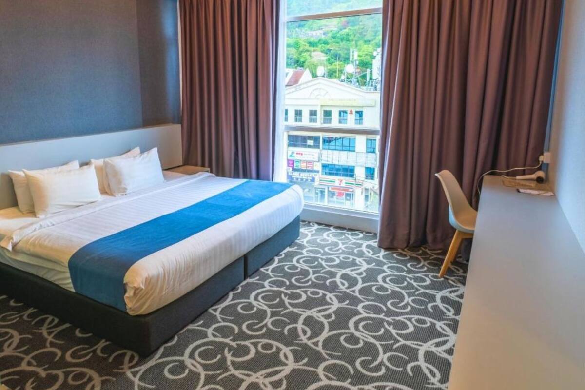 A hotel room with a bed, a view of a cityscape, and a patterned carpet.