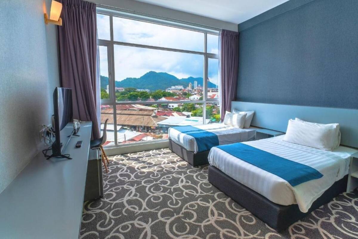 A hotel room with two beds, a large window with a view of a cityscape and mountains, a flat-screen TV, and a patterned carpet.