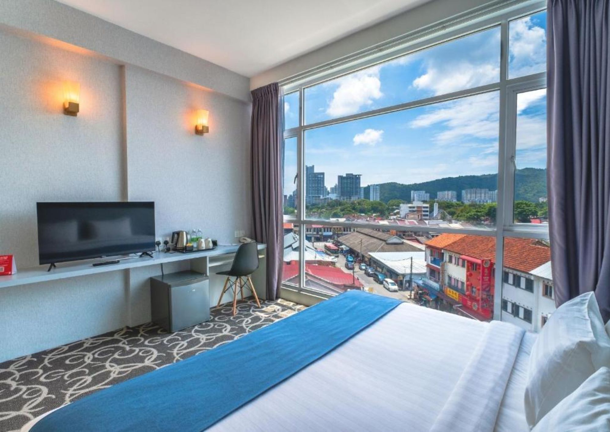 A hotel room with a bed, a desk, a TV, and a large window with a city view.