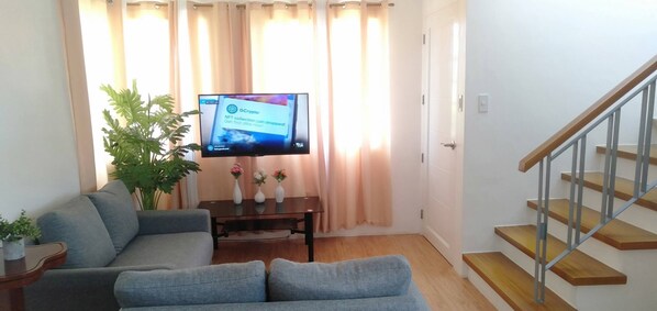 Una sala de estar con un sofá gris, una mesa de centro de madera, un televisor sobre un mueble, una planta en maceta y una escalera.