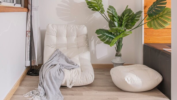 A cozy room with a white cushioned chair, a large green plant, and a wooden floor.