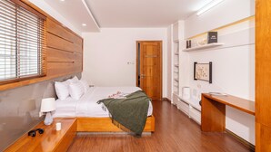 A modern hotel room with a wooden headboard, a bed with white linens, a bedside table with a lamp, and a desk with a chair.