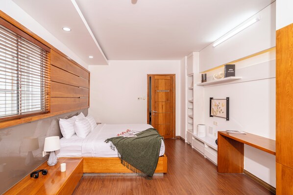 Une chambre d’hôtel moderne avec une tête de lit en bois, un lit recouvert de draps blancs, une table de chevet avec une lampe et un bureau avec une chaise.