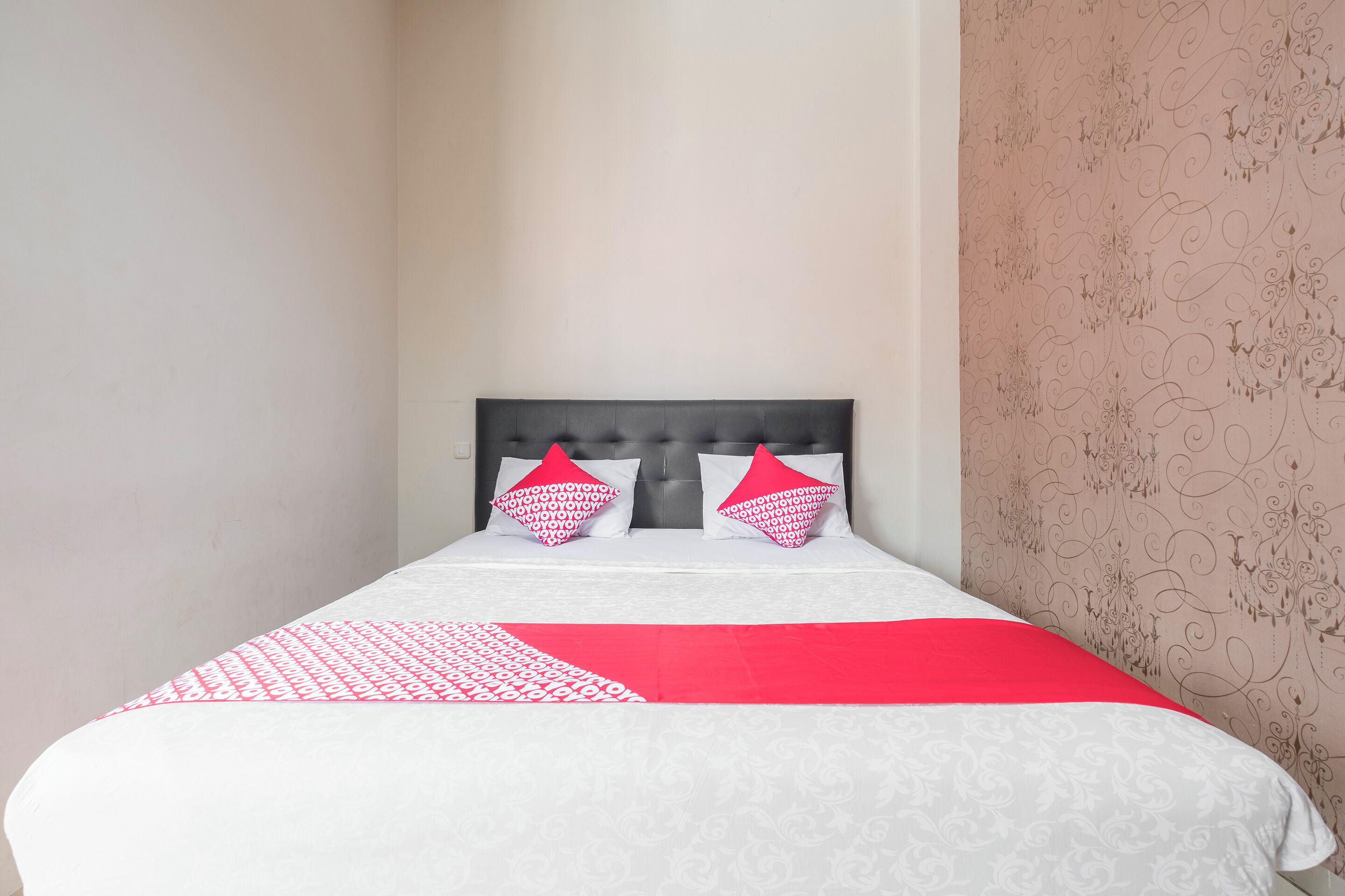 A neatly made bed with a patterned bedspread and two pillows, a red and white color scheme, and a decorative wall with a floral pattern.
