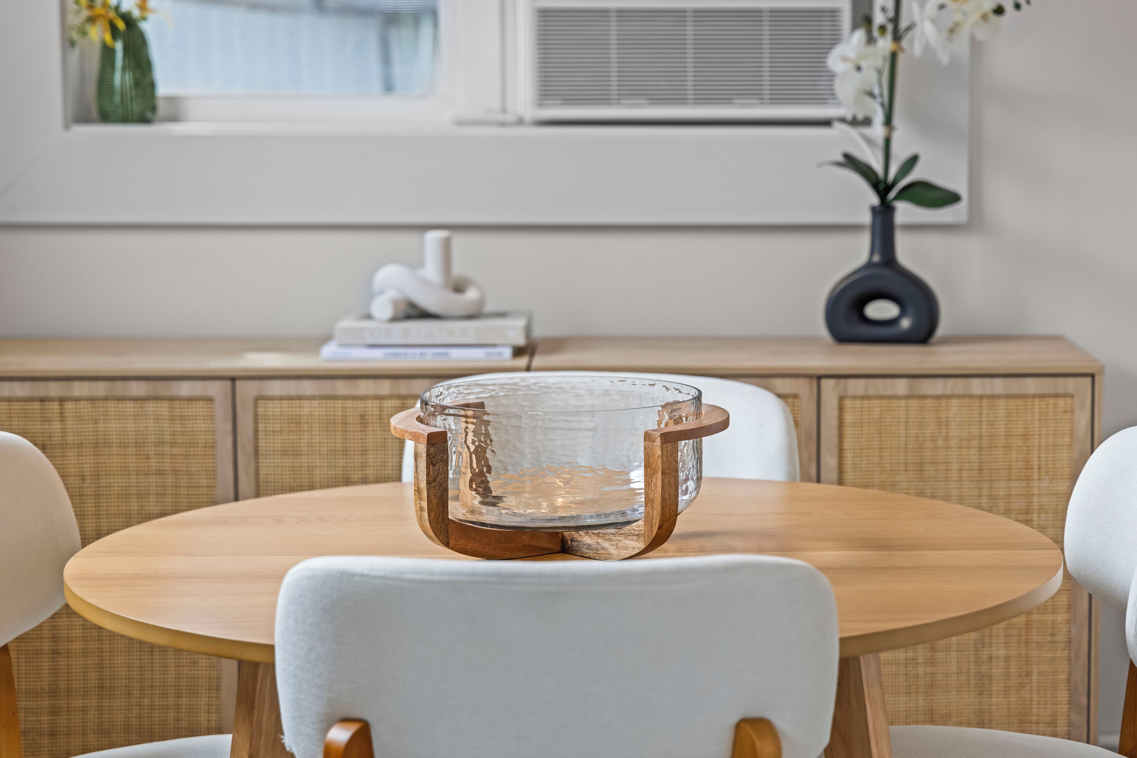 A round wooden table with a glass bowl centerpiece, surrounded by white chairs.