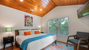 A bedroom with a wooden ceiling, a large bed with a blue and white bedspread, two bedside tables with lamps, a window with blinds, and a chair. - Under 1 mile to Dollywd, 5 mins Pkwy - 2 Kings/2 BA - Small fam or couples (Pigeon Forge)