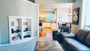 A modern living room with a wooden coffee table, a leather sofa, and a built-in bookshelf. A kitchen is visible in the background with a dining area. - Teak and Jordan Estates- Sleeps 28! (Albany)
