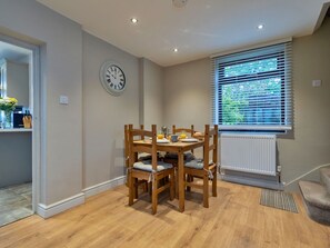 Un coin repas avec une table et des chaises en bois, une horloge au mur et une fenêtre avec des stores.