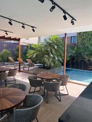 An outdoor patio area with round tables and wicker chairs, a swimming pool, and a covered section with ceiling lights. - Porto Esmeralda Apartment (Ipojuca)