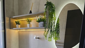 Un baño moderno con inodoro, lavamanos y espejo. Hay plantas en un estante sobre el lavamanos y una planta colgante a la derecha.