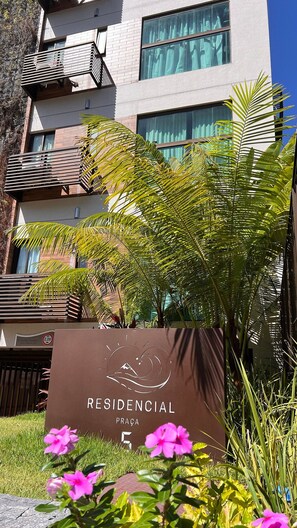 Letrero de 'RESIDENCIAL PRACA' con diseño de montaña y ola, rodeado de follaje y flores.