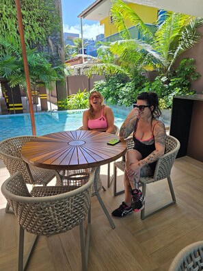 Two people seated at a round wooden table with wicker chairs, by a poolside area with a view of a building and greenery in the background. - Porto Esmeralda Apartment (Ipojuca)