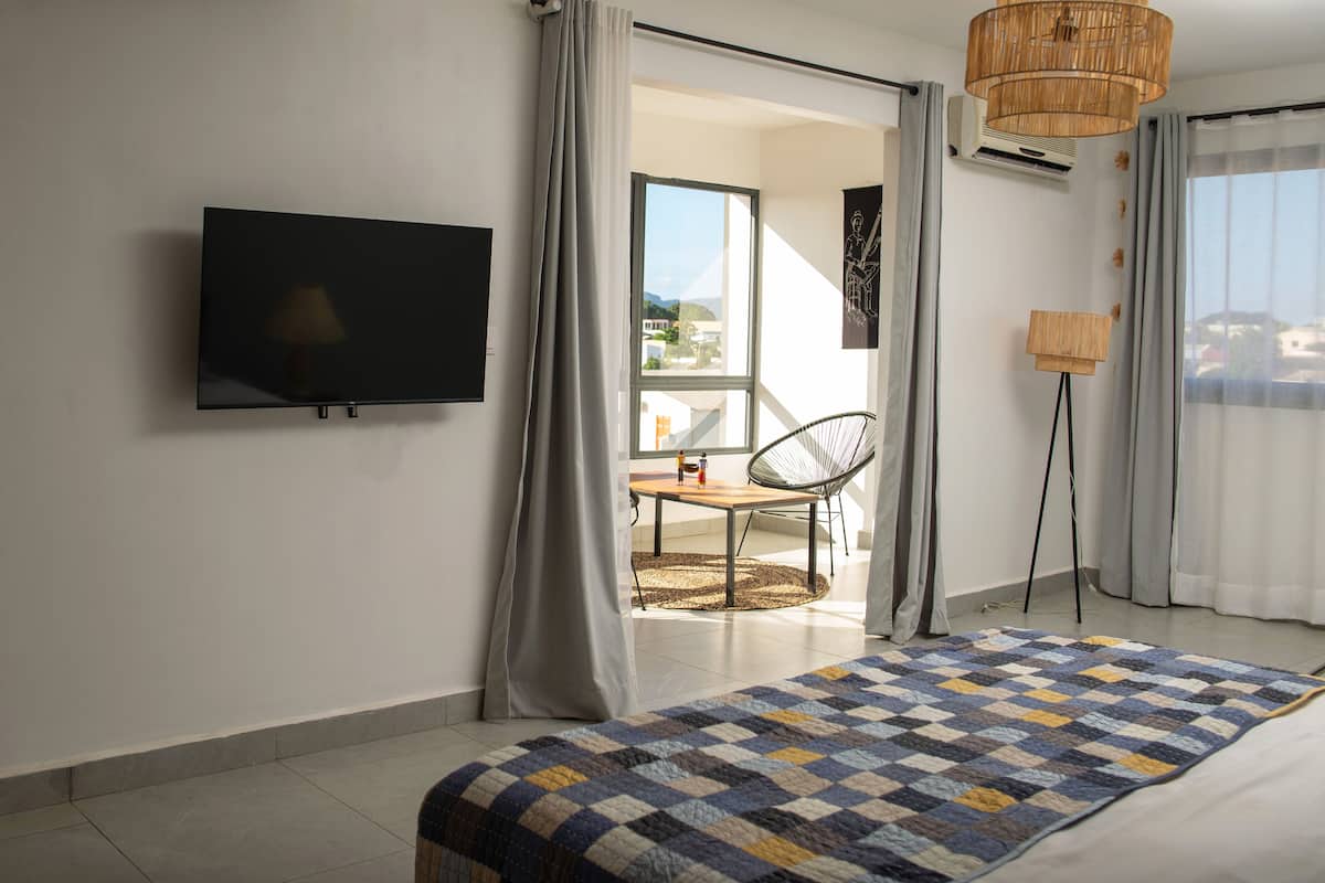 Une chambre moderne avec un grand lit, un téléviseur à écran plat fixé au mur, un coin repas avec une table et des chaises, et une vue dégagée sur l’extérieur grâce à une grande fenêtre.