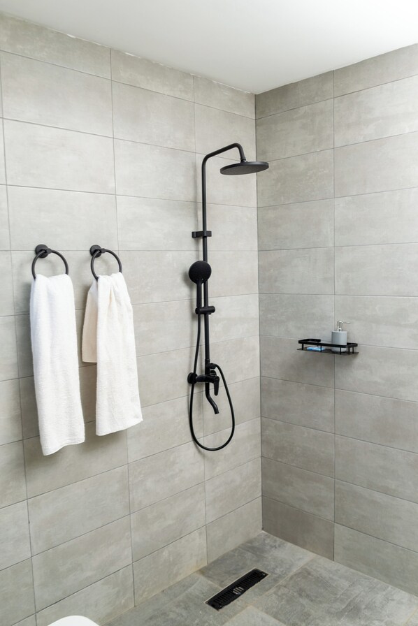 Un baño moderno con azulejos grises, un cabezal de ducha negro y dos toallas blancas colgadas en ganchos.
