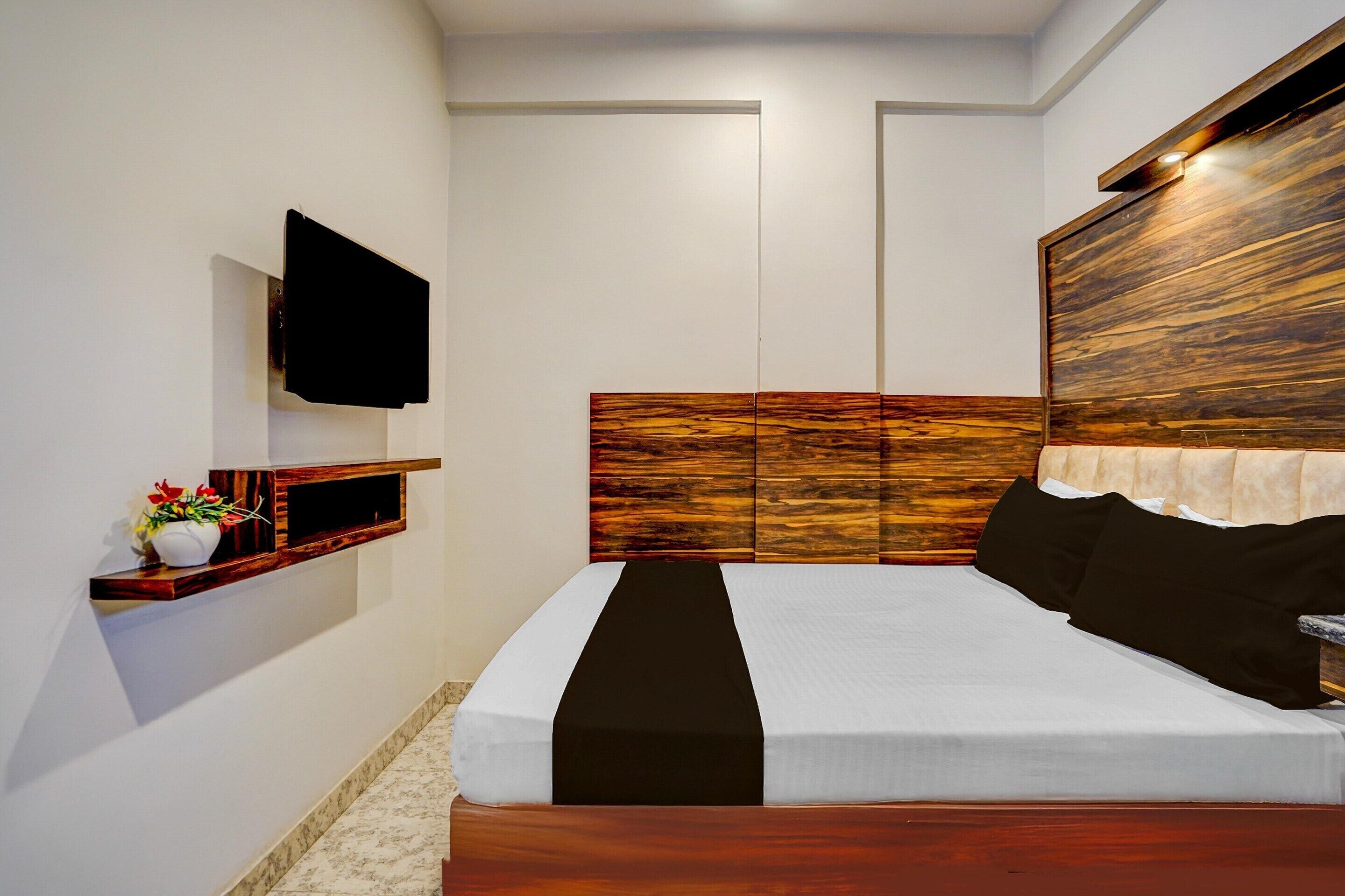 Un dormitorio con cabecera de madera, televisor de pantalla plana montado en la pared, mesita de noche con un jarrón de flores y cama con colcha blanco y negro.