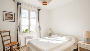 A bedroom with a bed, a chair, a bedside table, a lamp, and a window with curtains.
