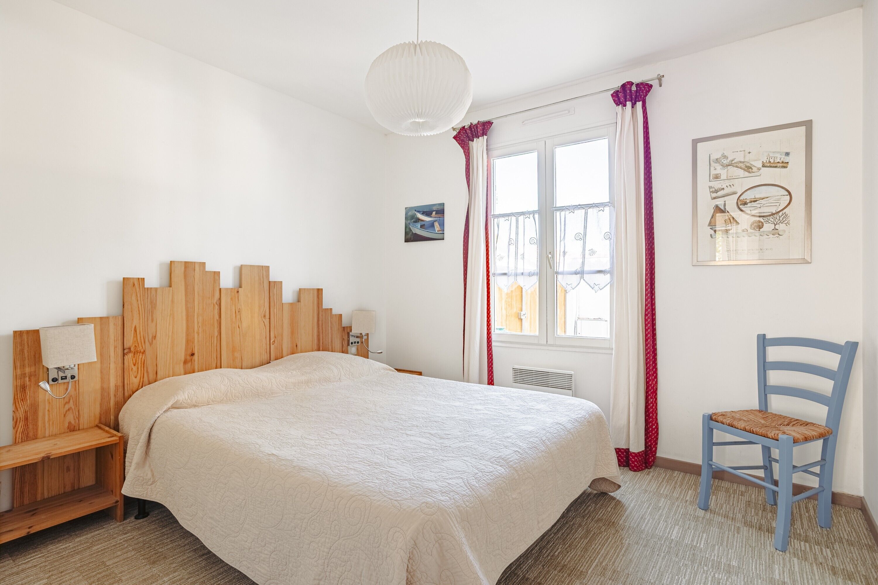 Een slaapkamer met een houten hoofdbord, een bed met een beige dekbedovertrek, een stoel, een raam met gordijnen en een wandlamp.