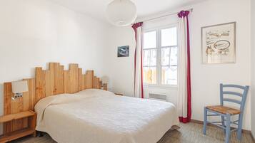 Een slaapkamer met een houten hoofdbord, een bed met een beige dekbedovertrek, een stoel, een raam met gordijnen en een wandlamp.