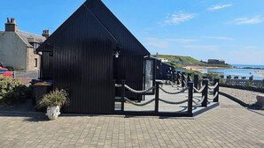 A black wooden building with a triangular roof, a paved walkway leading to it, and a view of the beach and sea in the background. - 2 Bed Cottage-sleeps 4-harbour & Sea View (Peterhead)