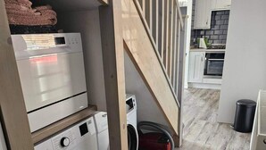 A laundry area with stacked dryers and a washing machine, a vacuum cleaner, and a staircase leading to a kitchen area with a stove and oven. - 2 Bed Cottage-sleeps 4-harbour & Sea View (Peterhead)