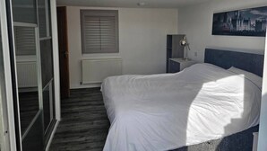 A bedroom with a bed, a radiator, a window with blinds, a desk with a lamp, and a painting on the wall.