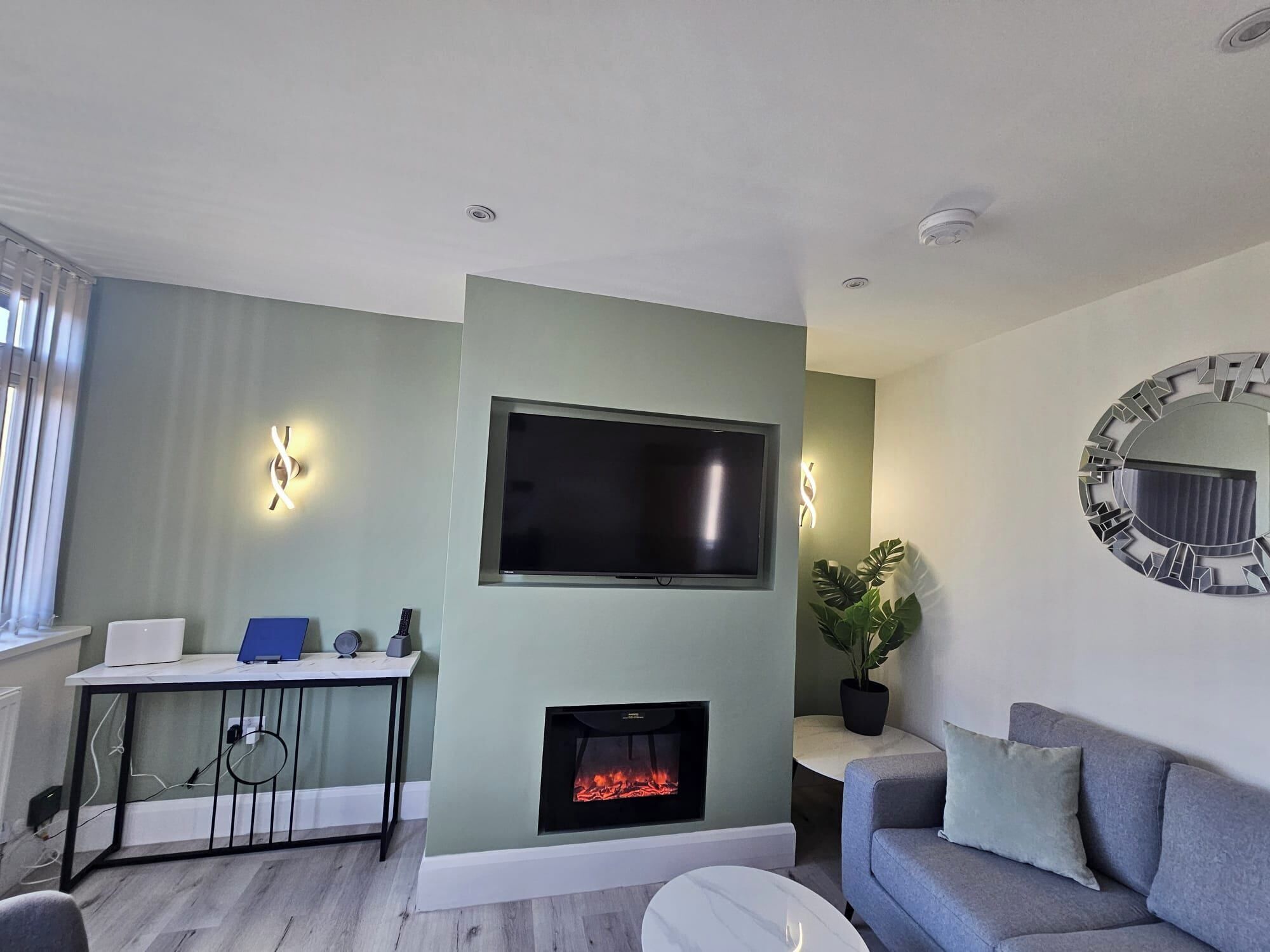 A modern living room with a flat-screen TV mounted above a fireplace, a sofa, a small table with a laptop and a speaker, and decorative wall lights.