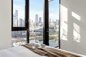 A hotel room with a large window offering a city view, a bed with a white comforter, and two white cups on a tray.
