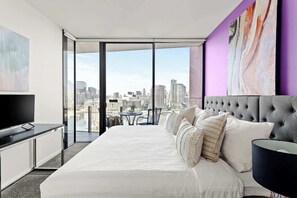 A modern hotel room with a large bed, a flat-screen TV, a dining area, and a city view through floor-to-ceiling windows.