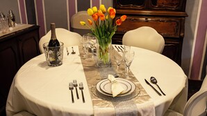 Une table ronde, dressée pour le dîner, recouverte d’une nappe blanche, avec une bouteille de champagne, un vase rempli de tulipes oranges et divers couverts.