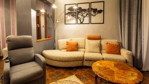 Un salon avec un fauteuil gris, un canapé beige agrémenté de coussins orange, une table basse en bois et des œuvres d’art murales représentant des arbres.