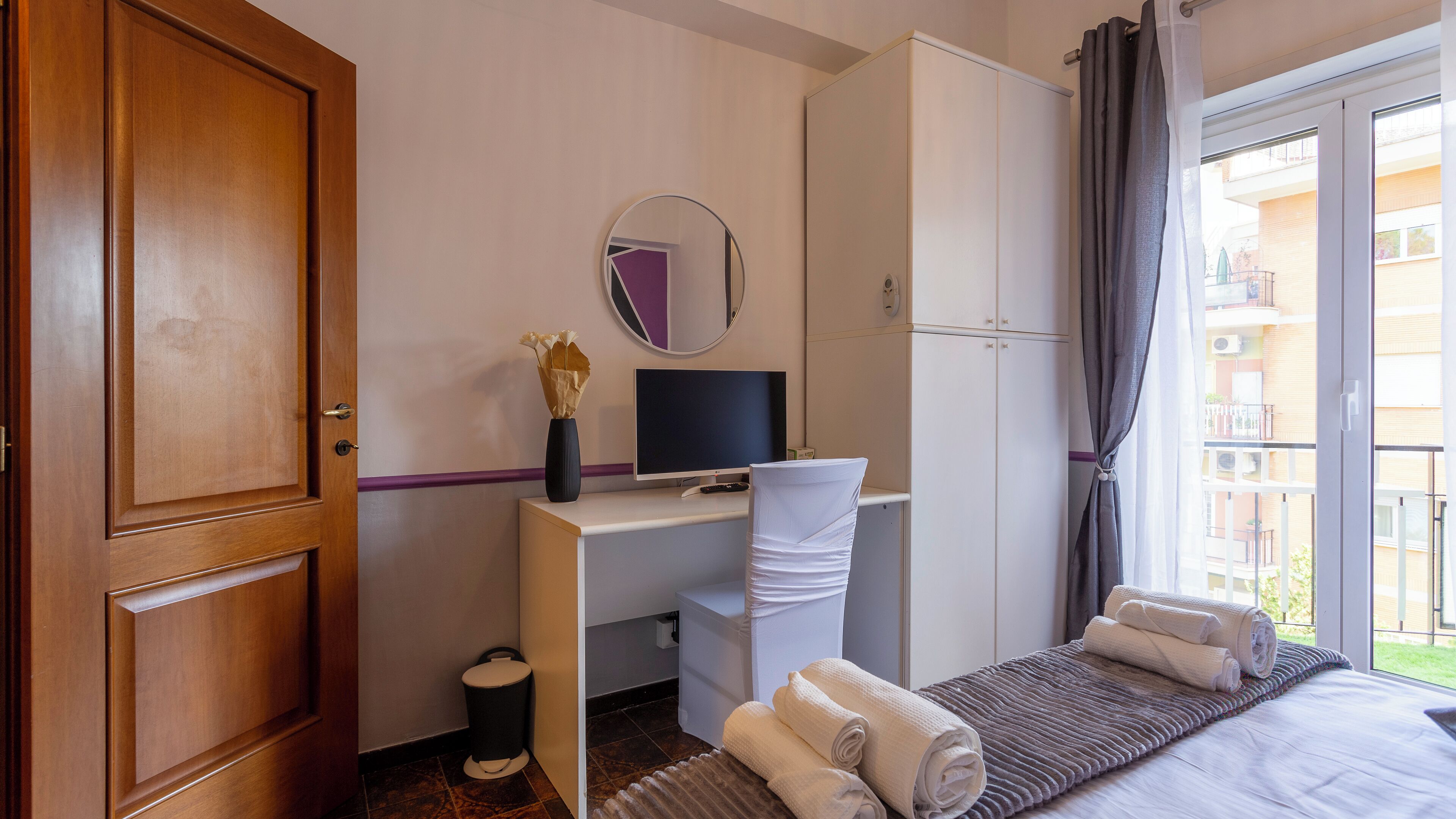 Habitació d'hotel amb una porta de fusta, un escriptori amb un ordinador, un mirall, un llit amb tovalloles i vista al balcó exterior.