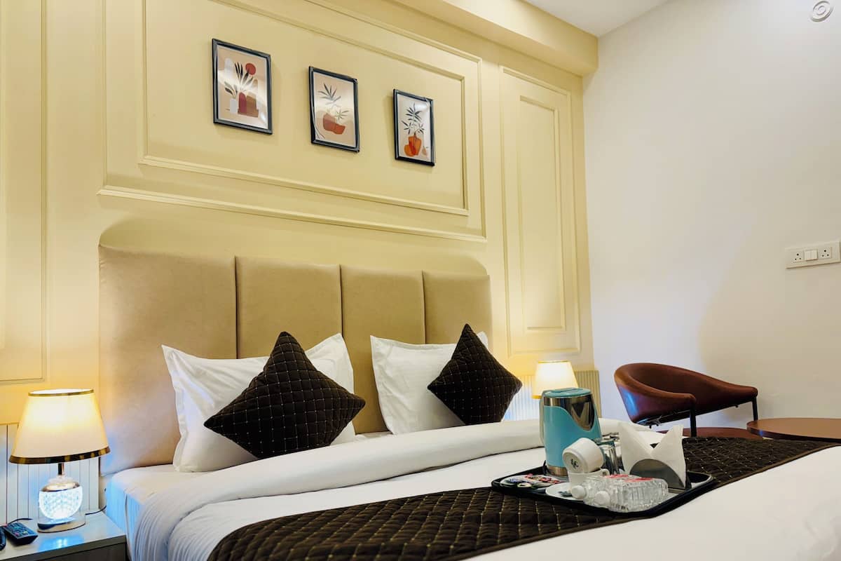 Una habitación de hotel con cama, mesita de noche, lámpara, sillón y una obra de arte colgada en la pared.