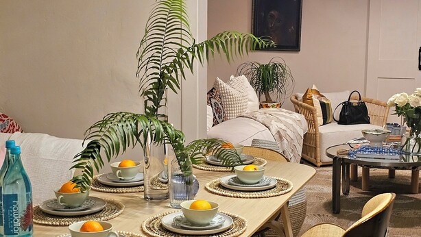 Een eetzaal met een lange tafel gedekt voor zes personen, omringd door stoelen. Er liggen geweven placemats, een vaas met een plant en staat een fles water op de tafel.