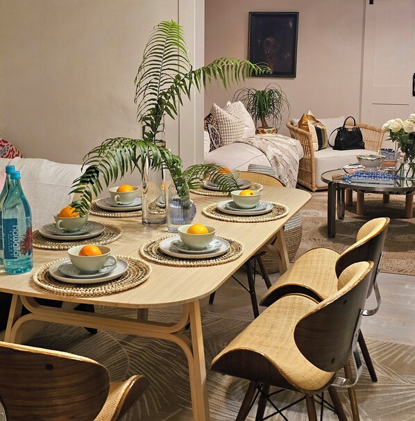 A dining room with a long table set for six, surrounded by chairs. There are woven placemats, a vase with a plant, and a bottle of water on the table.