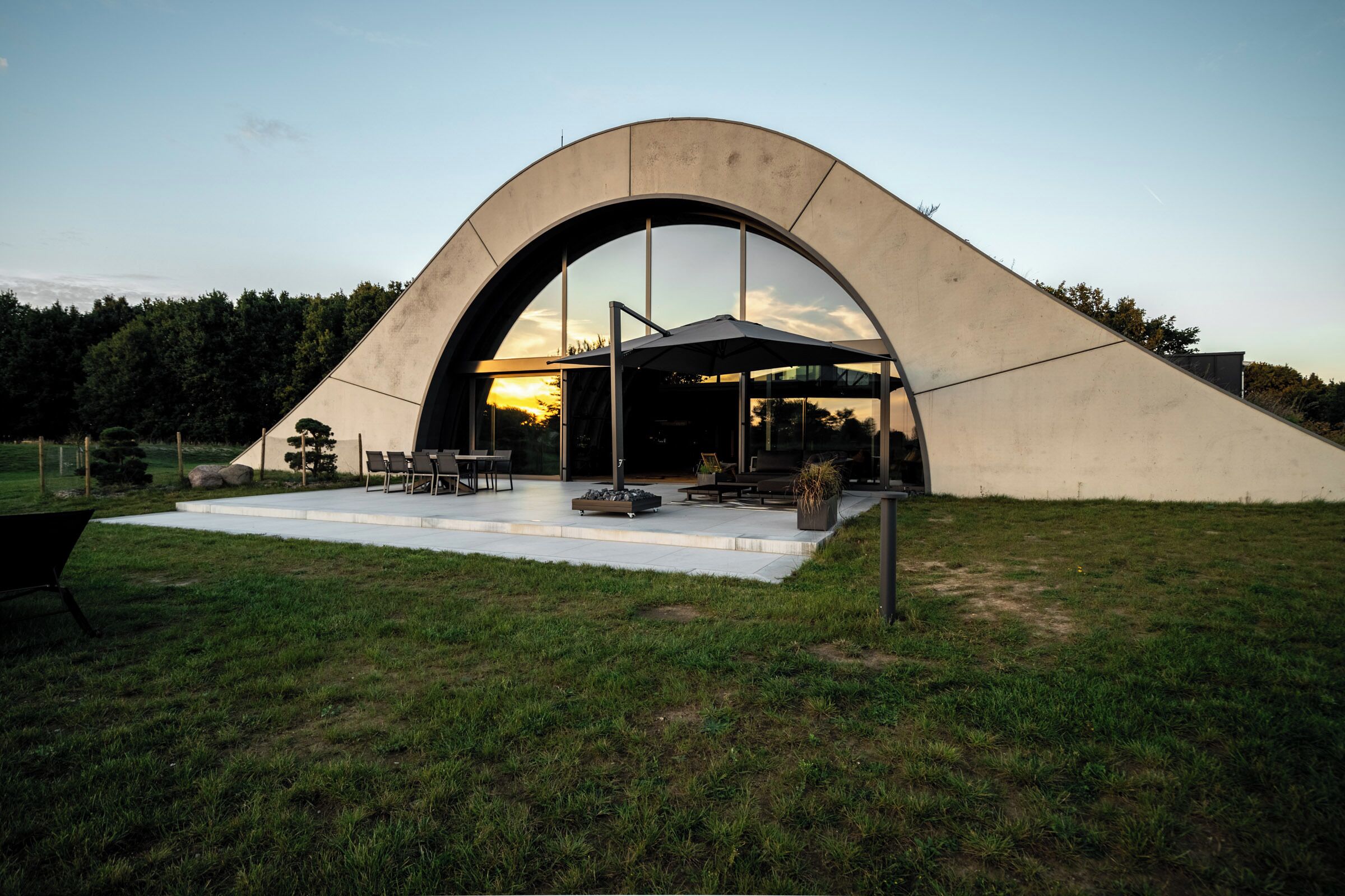 A modern building with a curved roof and large windows, surrounded by a grassy area with outdoor furniture.