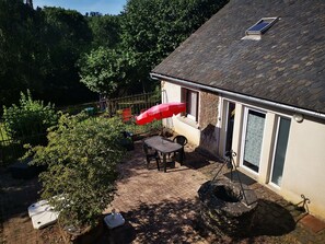 Ein Haus mit Ziegeldach, einer Terrasse mit Tisch und Stühlen, einem roten Sonnenschirm und einem Garten mit Büschen und einem Zaun.