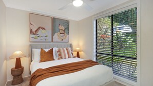 Une chambre à coucher comprenant un lit, deux tables de chevet, un ventilateur de plafond, une fenêtre avec des stores et deux œuvres d’art accrochées au mur.