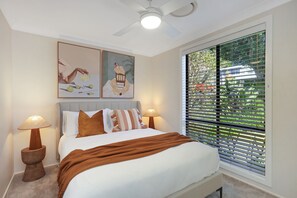 Un dormitorio con cama, dos mesitas de noche, ventilador de techo, ventana con persianas y dos obras de arte colgadas en la pared.