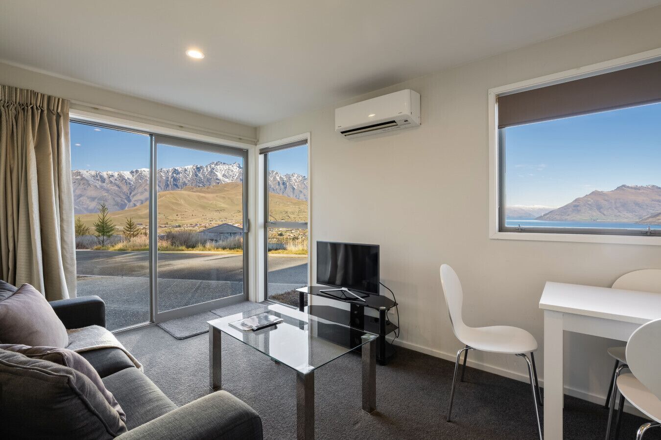 Une pièce moderne dotée d’une table en verre, d’un canapé, d’un téléviseur et d’un bureau. Elle offre de grandes fenêtres avec vue sur les montagnes.