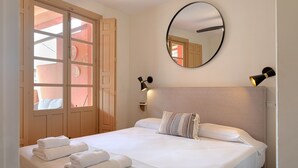 Habitación de hotel con cama, espejo redondo, dos lámparas fijadas a la pared y vistas a través de una puerta con ventana.